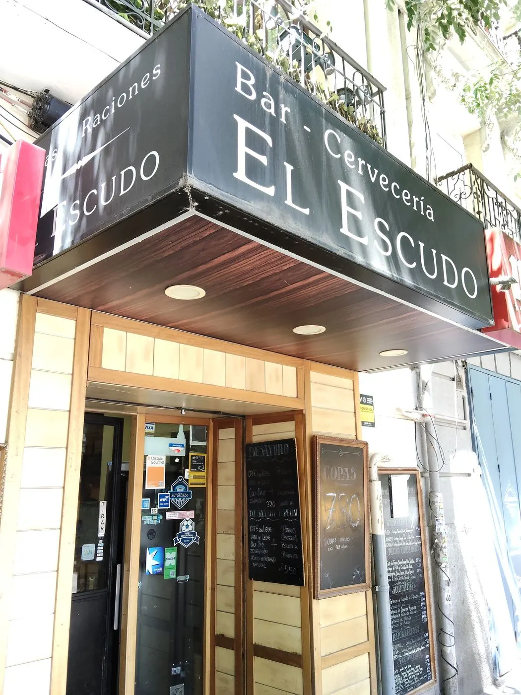 Entrada del bar con el rótulo Bar - Cervecería El Escudo, el tejadillo de madera y las pizarras de la puerta